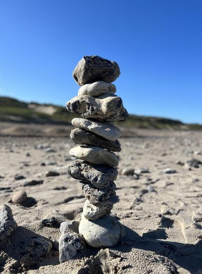 Stack of various sized and shaped rocks balanced vertically on a sandy beach, with small, scattered stones around and dune landscape in the background under a clear blue sky.