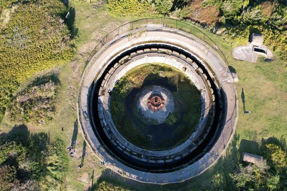 Aerial view of a large circular abandoned structure surrounded by grass and trees. The structure has concentric concrete layers leading down to a central pit with a rusted metal object. A path leads to the structure, and one side has small outbuildings.