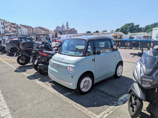 Photo from the front left of a Fiat Topolino quadricycle parked amongst motor scooters.