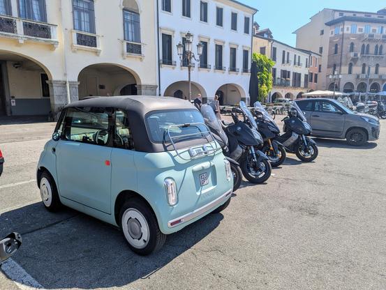 Photo from the rear left of a Fiat Topolino quadricycle parked amongst motor scooters.