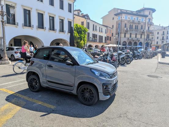 Photo from the front right of a Ligier JS50 microcar parked amongst motor scooters.
