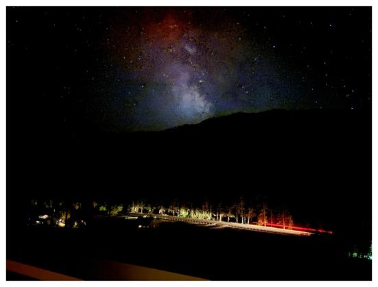 Landscape-orientation photo of the Milky Way rising in the middle of a starry sky, above the black silhouette of a tree-covered hill. It’s tinged red at the top and a brighter blue-white closer to the hilltop. At the bottom of the hill, a band of light — created by vehicle headlights captured in long exposure — illuminates a strip of highway and the bottoms of trees. A streak of red taillights runs parallel to the highway at right frame. Some house lights shine at lower left, on the near side of the highway. At the bottom of the frame in the extreme foreground is a glimpse of a deck guardrail.