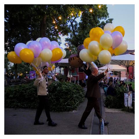Two men about 5m away, carrying lots of balloons in various colors across a public park, apparently going to a party somewhere. In the surrounding there are dark green trees and bushes, and people sitting around in a public park. (made by hand)