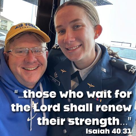 A smiling man wearing glasses and a blue hoodie poses for a selfie with a young woman in a military uniform. They appear happy, and the image features a quote from Isaiah 40:31.
