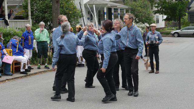 Six sword dancers with blue shirts and black trousers dancing in a street. They are in a circle, clashing longswords in the center. A musician plays a concertina. There is a small dog tethered to the musician. Behind them is an audience including a number of morris dancers.