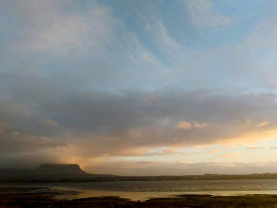 A darkening landscape on a rainy day. The sun has set and the clouds are breaking to reveal the evening sky. In the foreground, an expansive mudflat landscape with a large expanse of shallow water surrounded by green meadows and pastures can be recognised. On the horizon to the left, the iconic silhouette of Table Mountain Benbulben stands out against the atmospheric sky.

Eine dunkel werdende Landschaft an einem regnerischen Tag. Der Sonnenuntergang hat stattgefunden und die Wolken reißen auf und geben den Blick auf den  abendlichen Himmel frei. Im Vordergrund ist eine flache, ausgedehnte Wattlandschaft mit einer von grünen Wiesen und Weiden umgebenen großen flachen Wasserfläche zu erkennen. Am Horizont links zeichnet sich die ikonische Silhouette des Tafelbergs Benbulben vor dem stimmungsvollen Himmel ab.