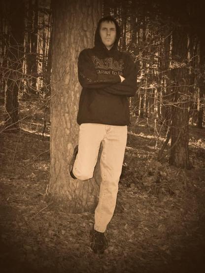 Thomas steht mit verschränkten Armen an einen Baum angelehnt und Blickt in die Kamera. Er trägt einen schwarzen Hoodie und weiße Jeans. Das Bild is sepia gefärbt