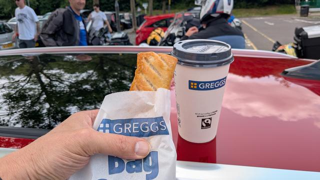 A one way cup of coffee with a Greggs label on it and a hand holding a paper bag with a sausage roll half out of it.