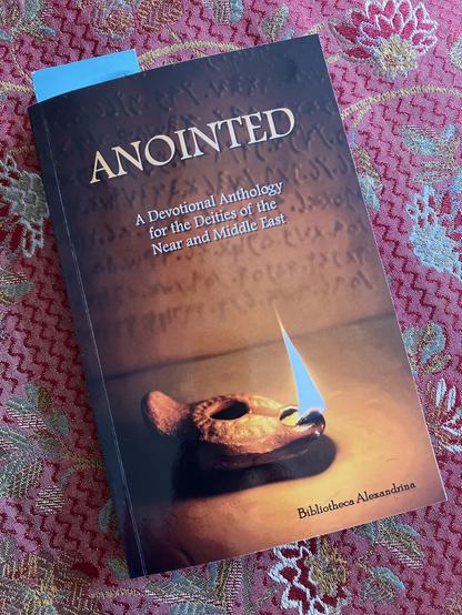 A paperback copy of Anointed: A Devotional Anthology for the Deities of the Near and Middle East, lying on a red jacquard fabric surface. The book cover includes the image of an ancient-style oil lamp with a burning wick. The top of a blue bookmark is visible at the upper edge of the book.