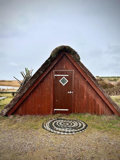 A triangular wooden hut with a moss-covered thatched roof sits in a grassy field. The hut features a red-painted wooden door with a white diamond-shaped window and a white upward arrow decoration. Adjacent to the hut, a pebble spiral pattern decorates the ground. A muted, overcast sky and faint outlines of distant vegetation complete the scene.