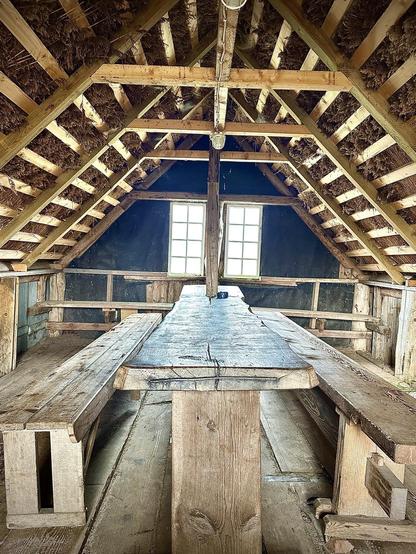 Interior of a rustic, wooden attic with a peaked roof supported by a central beam, flanked by two windows. The roof structure features exposed wooden beams and thatch insulation. A long wooden table runs down the center, accompanied by simple, bench-style seating. The floor is constructed from wide wooden planks.