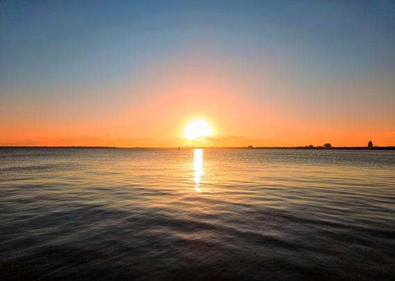 Sunset over the Saint Johns River. Warm colors form a halo over the firey yellow sun as it hits the horizon across a vast river, casting a beam of golden light over the soft waves in the otherwise quiet blue river waters.