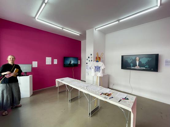 An art exhibition space featuring a bright pink wall, a woman holding a notebook, two screens displaying video content, and a table with printed materials. A T-shirt with a ribcage design is displayed on a stand. Various papers are spread across the table.