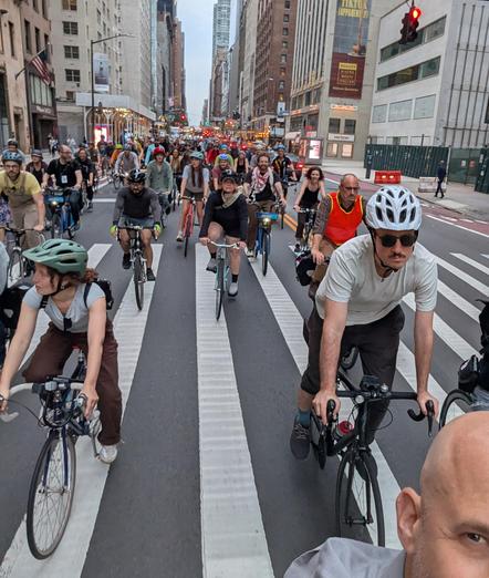 Cyclist proceed East on 57th Street in New York City on May 30 2025. Riders are looking forward while crossing a wide crosswalk. The photo is taken with a selfie camera looking west and a portion of the photographer's face is visible in the lower right corner