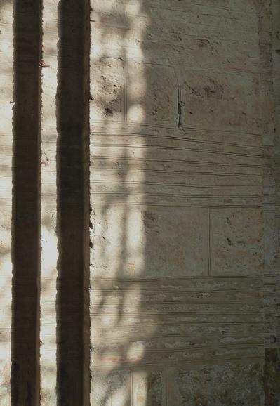Close up photograph of sunlight projected on the light brown surface of a medieval church pillar, casting intricate shadows and highlighting the texture and the lines formed by the stacking of the old cut stones.