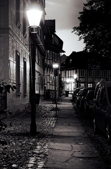 Narrow cobbled street at night, lined with half-timbered buildings on the left and parked cars on the right. Tall streetlights illuminate the path, casting shadows on the ground. Tree branches partially visible in the upper right corner. The scene is in black and white, creating a contrast between light and shadow.

Schmale, gepflasterte Straße bei Nacht, gesäumt von Fachwerkhäusern links und parkenden Autos rechts. Hohe Straßenlaternen beleuchten den Weg und werfen Schatten auf den Boden. In der oberen rechten Ecke sind teilweise Äste sichtbar. Die Szene ist in Schwarzweiß gehalten, wodurch ein Kontrast zwischen Licht und Schatten entsteht.
