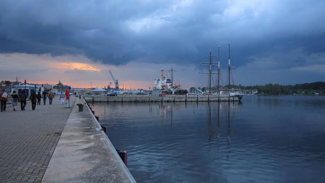 A wide shot shows a paved promenade running along a still body of water at dusk. Several people are walking along the promenade. Moored in the water are several boats and ships, including a large sailing ship with multiple masts. Across the water, there's a view of an industrial port area with cranes and various buildings, all under a dramatic, cloudy sky with a hint of orange sunset on the left.