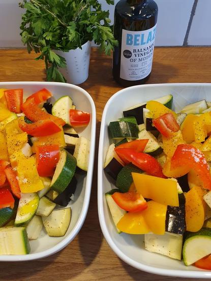 Two white dishes filled with colorful chopped vegetables, including zucchini, eggplant, and bell peppers in red and yellow. A small pot of fresh parsley and thyme is positioned nearby, along with a bottle of balsamic vinegar.