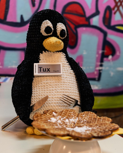 Ein gehäkelter Tux (mit Namenschild, Pronomen: Pingu/ihn) hält Messer und Gabel mit den Flossen und sitzt vor einer Waffel.