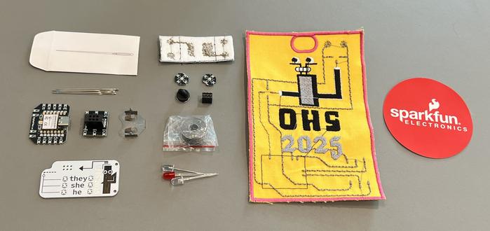 Unevenly knolled objects from the badge kit: various needles, the microcontroller, the SAO with pronouns, some LEDs, a sewable coin cell battery holder, a roll of conductive thread, the badge itself, a SparkFun sticker, and some other bits & bobs.