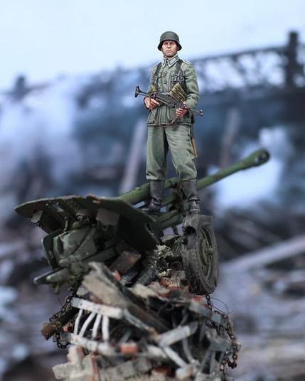 This is a strikingly detailed diorama or miniature scene, meticulously crafted to depict a dramatic moment in wartime history. The focus is on a lone soldier in World War II-era uniform standing atop a destroyed artillery piece, which is perched precariously on a pile of rubble. The soldier is rendered with incredible realism, his uniform crisp and accurate to historical details, featuring meticulously painted insignias and creases in the fabric. His expression is stoic yet determined, giving the scene an emotional gravity. He clutches a submachine gun—a German MP40—across his chest, a pose that exudes authority and readiness. His weathered boots and the slight wear on his helmet add a subtle touch of realism that suggests he has endured hardship.