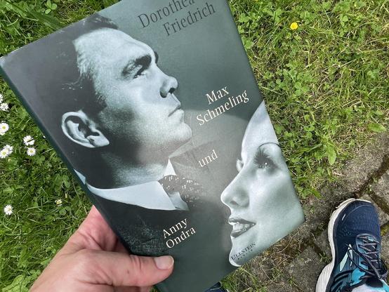 Foto eines Buches in meiner Hand mit dem Titel „Max Schmeling und Anny Ondra“, im Hintergrund angeschnitten Laufschuhe und Rasen