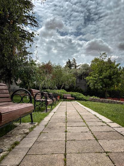 Photo that shows a pathway in a park. There are empty benches near the path. The grass and trees are green. Sky is cloudy.