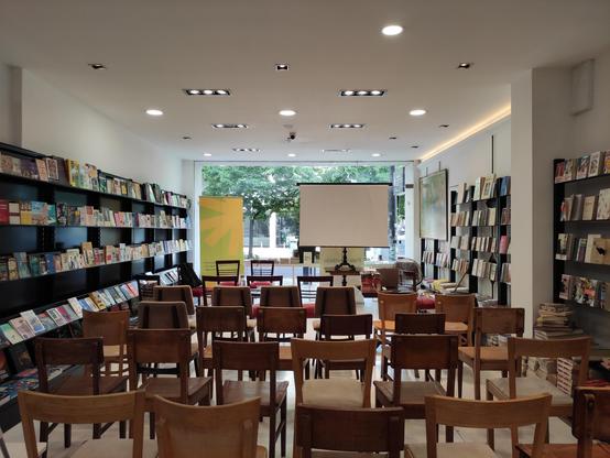 Foto da sala da livraria Castro e Silva, cheia de cadeiras, ladeada por estantes com livros e, ao fundo, a montra, onde está montada uma tela de projecção.
