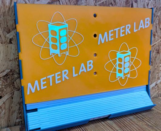 closeup eines in 3 farben 3D gedruckten und im realraum in FreeCAD gezeichneten Meterstab-Spender mit Metalab-Logo und Beschriftung "Meter Lab". Natürlich in Metlab Blau, Gelb und floureszierendem Weiß