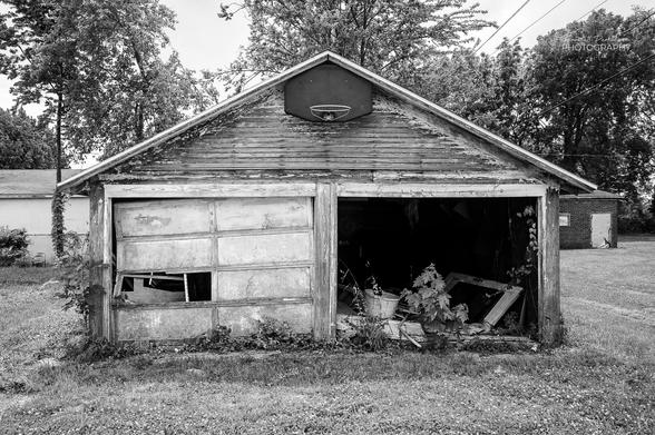 An old, abandoned two car garage with a basketball backboard attached at the peak of the roofline.  One garage door is missing and the other is off its rails on the left side with one broken panel.
