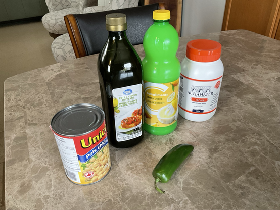 Containers of chickpeas olive oil lemon juice tahini and a whole green jalapeño pepper on a table.