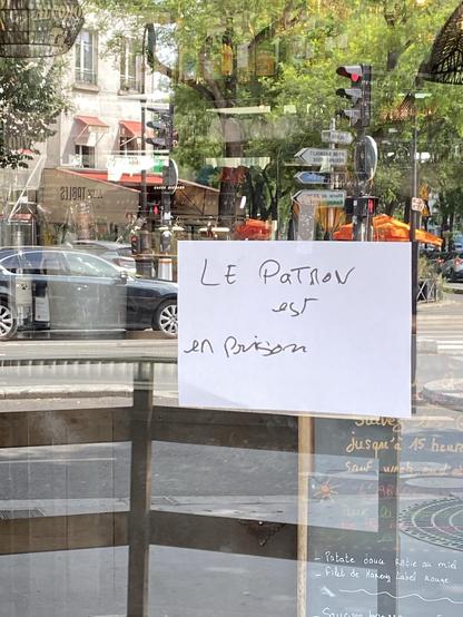 affiche blanche accroché à une porte en verre d’un restaurant, inscription: “Le Patron est en prison”; en reflection un carrefour de Paris