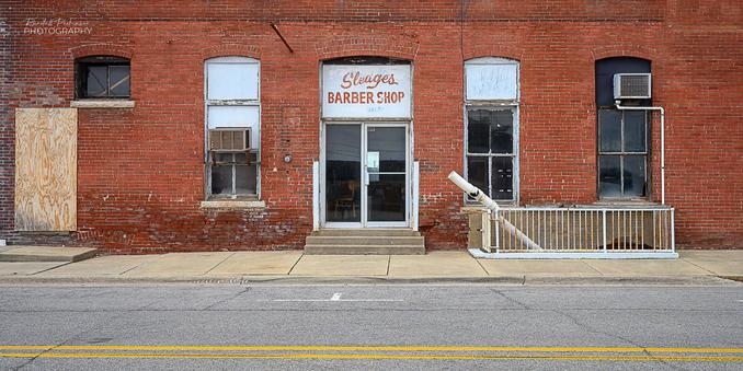 An old brick building with three tall windows and a double door that used to house a barber shop called Sleages Barber Shop.  A white fence blocks off a stairwell to a basement entrance.  A piece of 6" PVC pip with fittings lays in the stairwell.