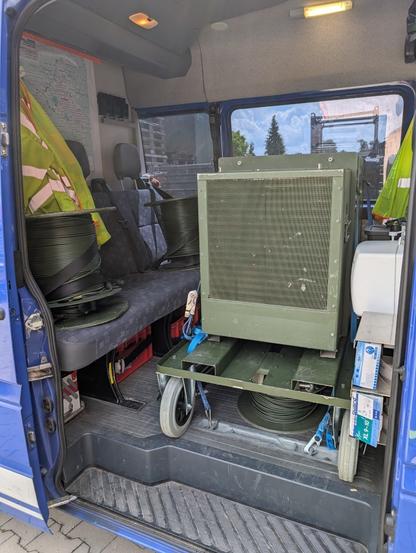 Das Feldrack steht im Inneren eines MB Sprinter, mit Spanngurten am Boden gesichert. Die Kabeltrommeln sind mit Anschnallgurten auf den Sitzen des Sprinters befestigt. Außerdem zu sehen sind Warnjacken und eine Hygienestation