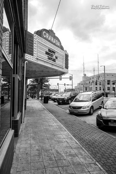 A black and white image looking along the length of a sidewalk with a large overhanging sign structure reading "Granada" for the Granada movie theater.  The text on the sign says "Happy Sweet 16 Zaylee".