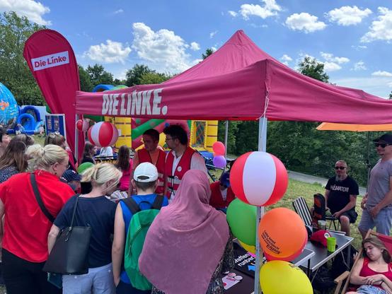 Das Bild zeigt eine Veranstaltung im Freien, bei der eine Gruppe von Menschen sich um einen roten Zeltstand versammelt hat, der mit dem Slogan "DIE LINKE" versehen ist. Der Zeltstand ist mit bunten Luftballons geschmückt, darunter rote und weiße sowie gelbe und orangefarbene. Im Hintergrund ist ein Inflatenturm zu sehen, der Teil eines Kinderbereichs ist. Einige Personen tragen rote Westen, was darauf hindeutet, dass sie möglicherweise Teil einer Organisation sind. Einige Besucher stehen und unterhalten sich, während andere sitzen. Die Atmosphäre wirkt freundlich und familienfreundlich. Der Himmel ist blau mit einigen weißen Wolken, und die Umgebung ist grün mit Bäumen.