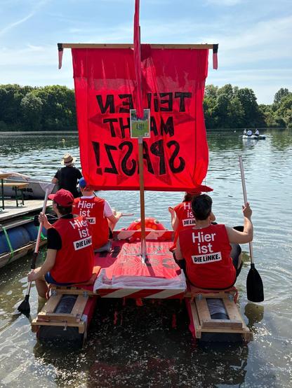 Das Bild zeigt eine Gruppe von Menschen, die in einem selbstgebauten, roten Ruderboot auf einem ruhigen Gewässer sitzen. Das Boot ist mit einem großen roten Segel ausgestattet, auf dem in schwarzen Buchstaben "TEILEN MACHT SPASZ!" steht. Die Personen tragen rote Westen mit der Aufschrift "Hier ist DIE LINKE." Sie paddeln mit Ruderstöcken, die sie in der Hand halten. Im Hintergrund sind weitere Boote und ein Ufer mit Bäumen zu sehen. Der Himmel ist blau mit einigen weißen Wolken. Die Szene wirkt entspannt und friedlich, während die Gruppe offensichtlich an einer politischen Veranstaltung teilnimmt.