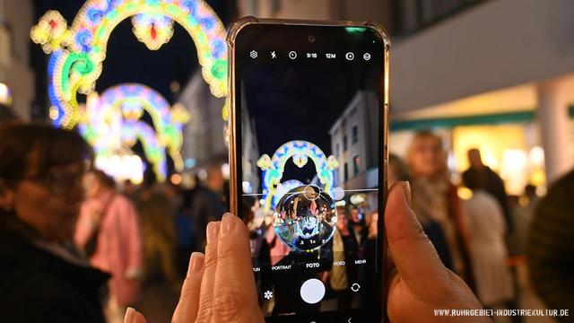 Ein Smartphone wird von zwei Händen gehalten und fotografiert eine gläserne Kugel vor bunter Straßenbeleuchtung auf einem nächtlichen Stadtfest. Im Hintergrund ist ein Lichterbogen mit floralen Mustern verschwommen zu erkennen, aufgenommen beim „Unna Festa Italiana“.