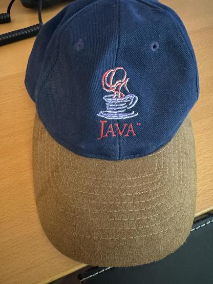 A top view of a ballcap, sitting on a desk.  The hat part is a dark blue, while the bill is a brown suede, with many equally-spaced sewed lines on it.

On the hat itself is a embroidered emblem for Java, showing the word in red, and the symbol of a white coffee cup and saucer, with red steam going up.

Got this for some event, can't remember where or when.
