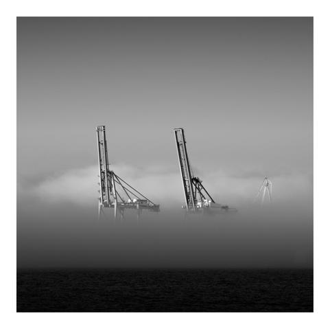 Three large industrial cranes emerge from a dense layer of fog above dark, calm water. The black-and-white image emphasizes stark contrasts and minimalism. The cranes appear ghostly and surreal, partially obscured by mist, evoking themes of impermanence and industrial isolation.