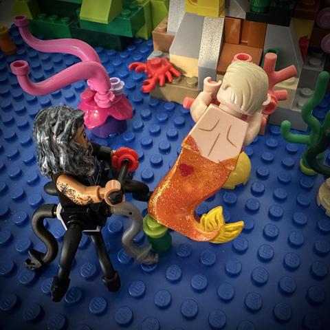 Lego MerKraken Ed holding a needle and red flower to apply a heart tattoo on Lego MerStede’s tail. Stede is facing away from the camera, holding onto a piece of coral.