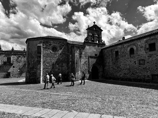 Una plaza empedrada, en blanco y negro, se encuentra en el centro de una antigua ciudad. En el medio de la plaza hay unas pocas personas caminando. A la izquierda hay un edificio de piedra más pequeño, y a la derecha, un edificio más grande, con una torre de campanario en el centro. El cielo está nublado. La imagen tiene una atmósfera tranquila y pacífica.