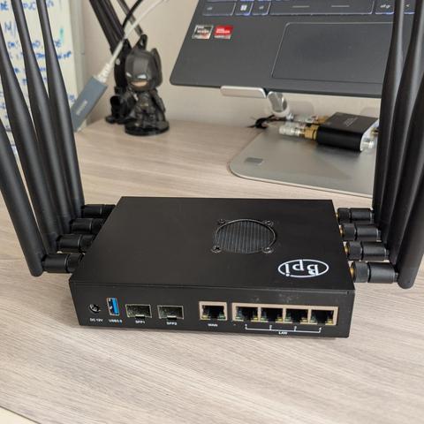 Imagen de un router negro con 8 antenas externas. El router está situado sobre una mesa de madera clara, con un portátil y una figura de Batman de juguete en el fondo. El router tiene varios puertos, incluyendo puertos USB, SFP, WAN y LAN, visibles en la parte trasera. El logotipo de la marca "Bpi" del router es visible en la parte superior.