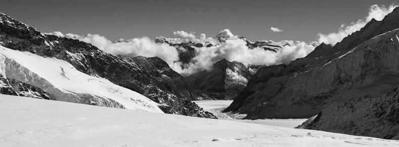 Snow-covered mountainous landscape in black and white. Foreground features a vast, snowy slope, leading to rugged, rocky peaks in the midground. Deep valleys and ridges are visible, with clouds gathering around some peaks in the background, under a clear sky.
