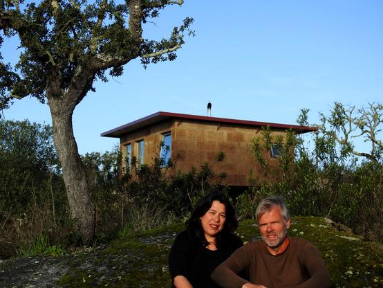 A couple, Tânia and Waldemar, sitting on a mossy granite rock and surrounded by holm oaks. Behind the reception of Bubulcus & Bolotas Camping, an eco-frindly building with cork façades.