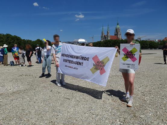 Bei schönstem Wetter halten zwei Menschen ein Transpi mit der Aufschrift "Menschenwürde verteidigen, AfD-Verbot jetzt" auf der Theresienwiese in München hoch. Im Hintergrund sieht man die Paulskirche.