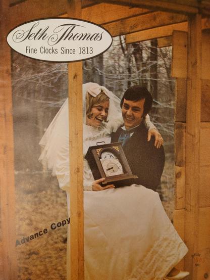 The cover of a Seth Thomas clock catalogue depicting a groom carrying his new wife over the threshold of their house. The wife is clutching a large shelf clock and both the man and the woman are laughing hysterically.