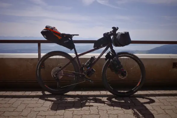 The Karate Monkey fully loaded with all the bags for the Trans Jura next week, leaning against a railing with the sunny blue sky and the Alps visible in the background.
