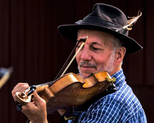 Ein Mann mit grauem Bart und einem Hut spielt die Geige. Der Blick ist auf die Saiten gerichtet, während der Bogen der Violine Klänge entlockt. Der Mann trägt ein blau-kariertes Hemd und steht vor einer dunkelroten Wand. 
. 
A man with a grey beard and a hat plays the violin. His eyes are fixed on the strings while the bow draws sounds from the violin. The man wears a blue chequered shirt and stands in front of a dark red wall. 
.