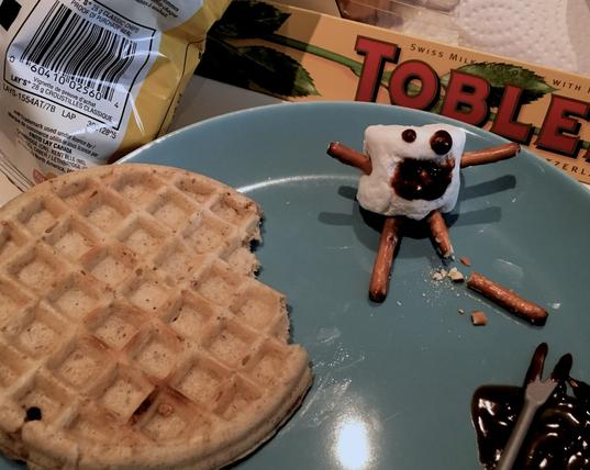 A waffle with a bite out of it sits on the side of a blue plate. Next to it are a smudge of chocolate and a marshmallow person with pretzel rods for arms and legs. One of it's legs has broken in half, there are dots of chocolate on it that make eyes and a mouth. The effect makes it look like the marshmallow person is screaming in pain because of it's broken pretzel leg. Next to the plate is a bag of chips and a box of Toblerone.